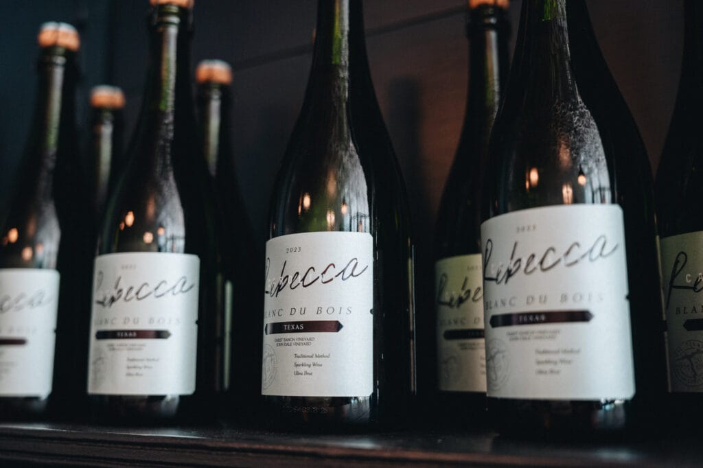 A row of Rebecca Caroline sparkling wine bottles lined up on a dark shelf