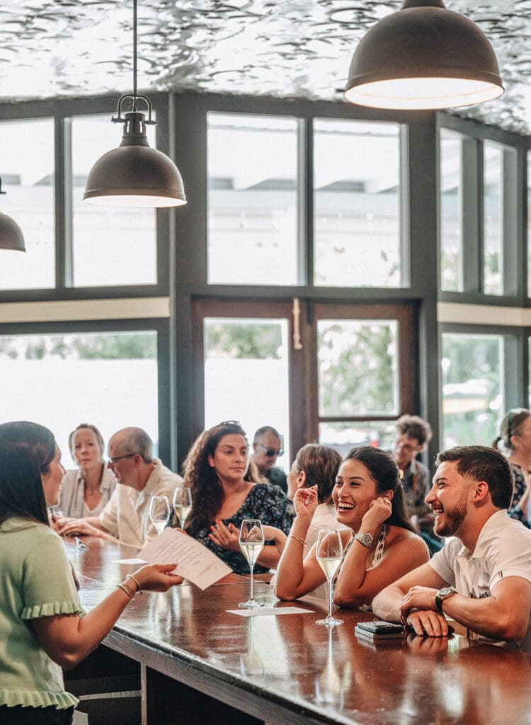 Guests seated along a communal table beside the tall windows of the tasting room