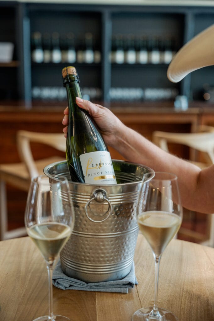 A bottle of Rebecca Caroline chilling in an ice bucket beside two flutes on a tasting room table